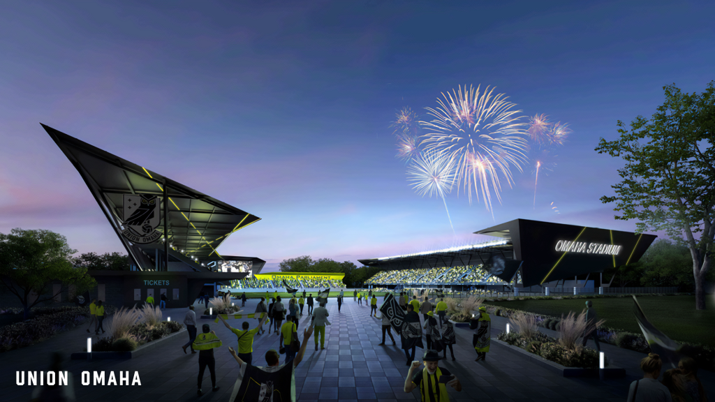 A ground-level view from outside Union Omaha's proposed new stadium, showing fans walking in with banners and flags as fireworks explode above the stadium.